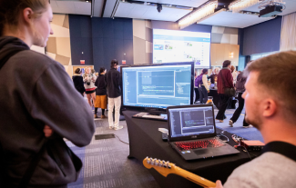 Two people looking at two computer screens. One person is holding a guitar.