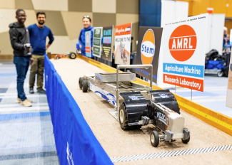 Wide shot of an RC tractor pull on a long table with three people in the distance holding the controller